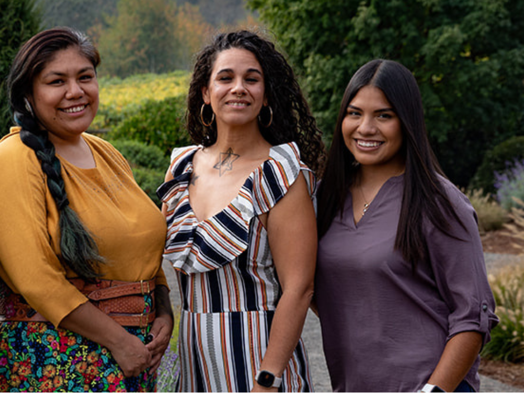 Three members of OCJ’s inaugural Community Cohort standing outdoors, smiling at the camera. One wears a yellow top, one a striped dress, and one a purple blouse.