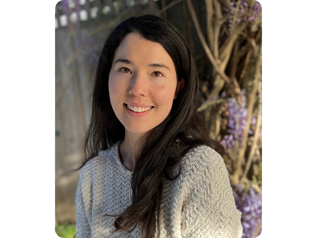 Headshot of woman with long dark hair in a sweater smiling at the camera with a background of wisteria blooms