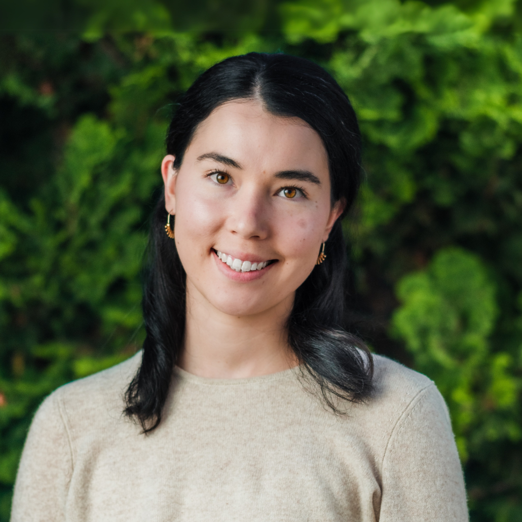 A woman with black hair, wearing a cream-colored sweater and gold earrings, smailing in an outdoor setting with a blurred background of trees and bushes.