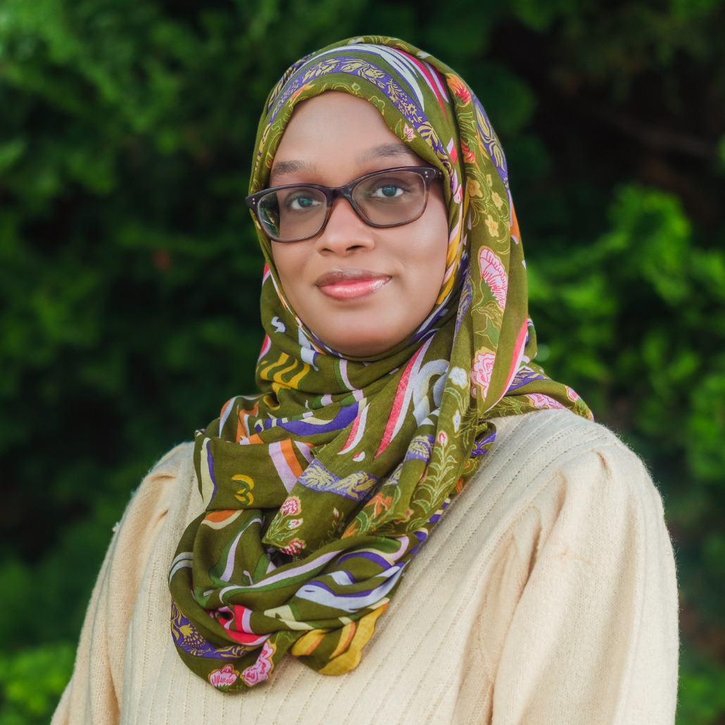 A woman with a yellow sweater and a green, purple, and pink scarf, wearing glasses, smiling in an outdoor setting with a blurred background of trees and bushes.