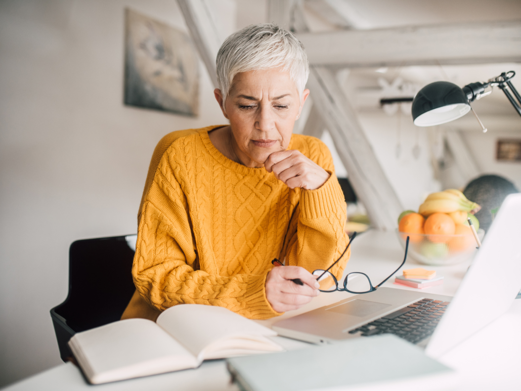 Woman with short white hair wearing a yellow sweater, sitting and looking at papers on a desk.