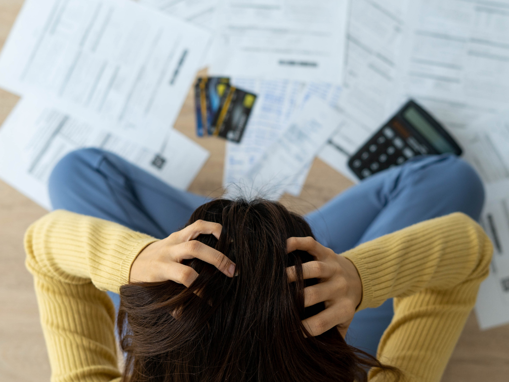 A person sitting on the floor with their head in their hands, surrounded by a pile of bills.