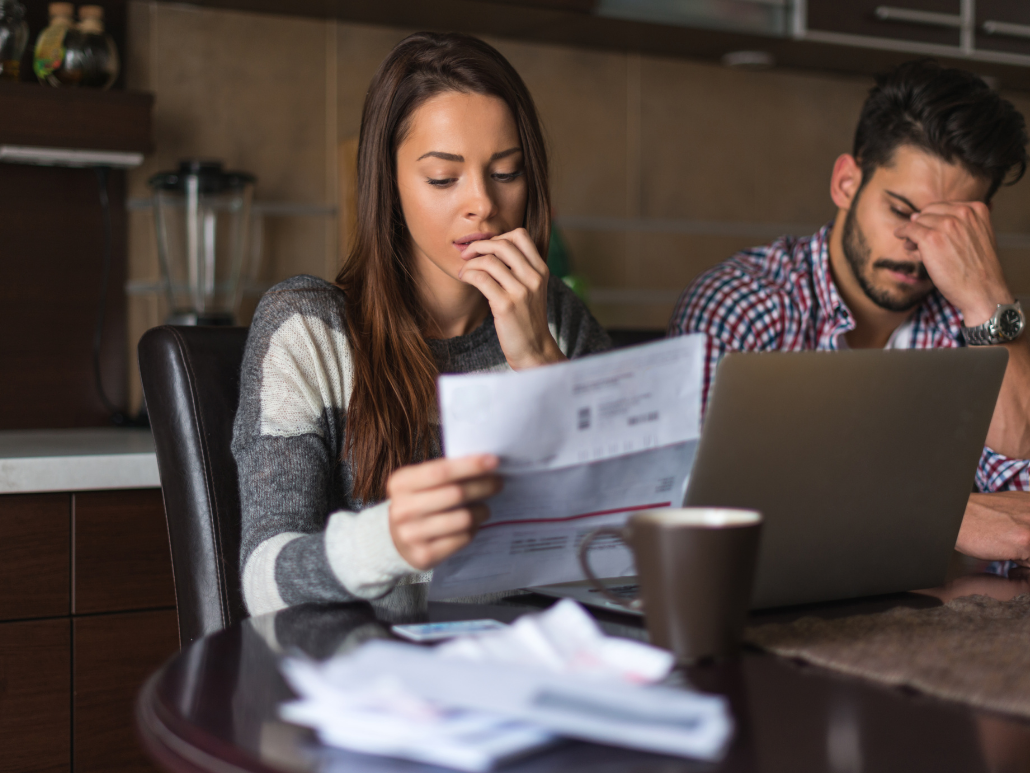  Two people are sitting at a table with a laptop and papers in front of them, looking concerned and upset. 