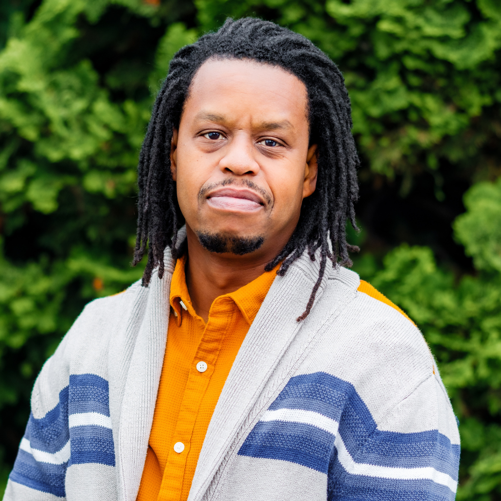 A man with dark brown dreadlocks, wearing a blue and white cardigan over an orange shirt, smiling, in an outdoor setting with a blurred background of trees and bushes.