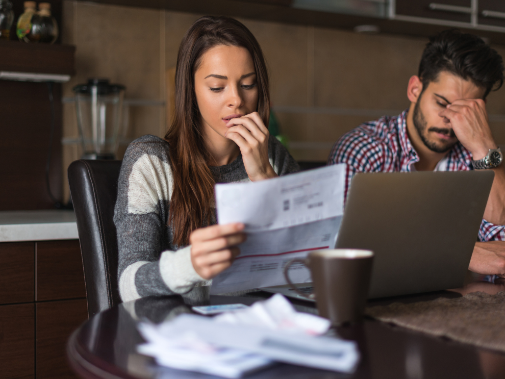 Two people are sitting at a table with a laptop and papers in front of them, looking concerned and upset.