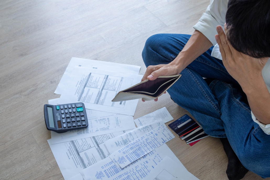 A person sitting on the floor holding their head, looking at a pile of papers and a calculator on the floor in front of them.