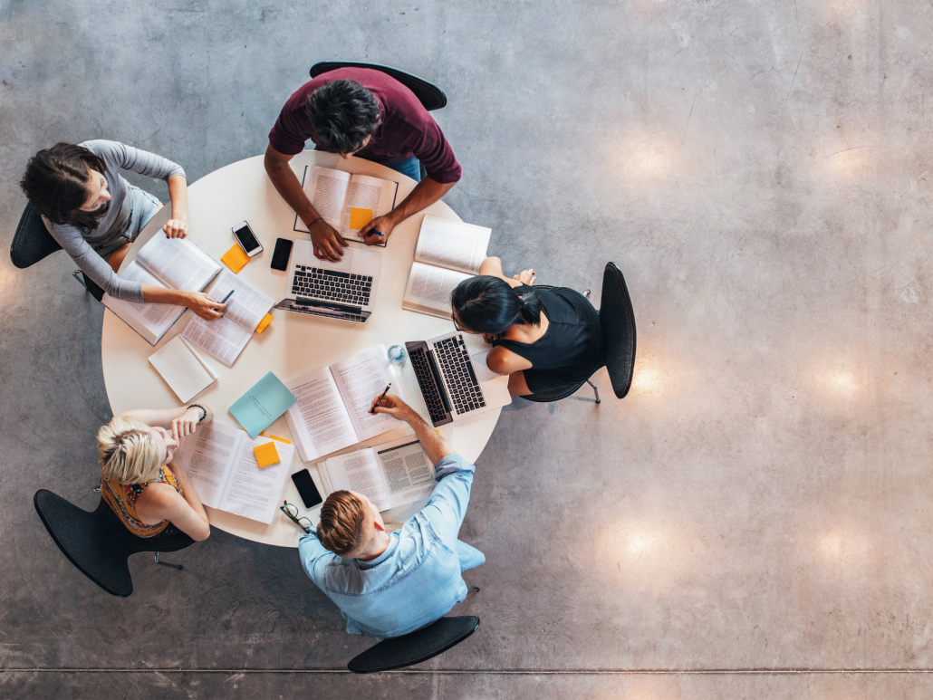 A birds eye shot of a group of people sitting around a table talking and working on computers.