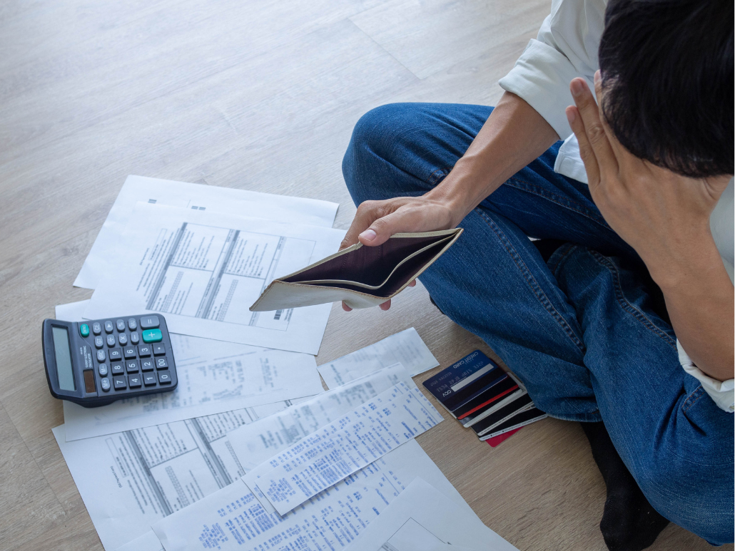 A person sitting on the floor in front of papers and credit cards. They are holding their head in one hand and an empty wallet in the other.