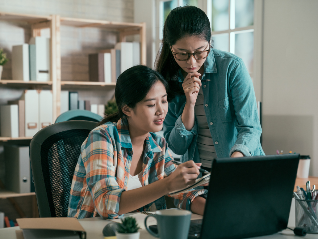 Two women looking at a computer. One woman is sitting down at and is pointing to a notebook with a pen. The other woman is standing up.
