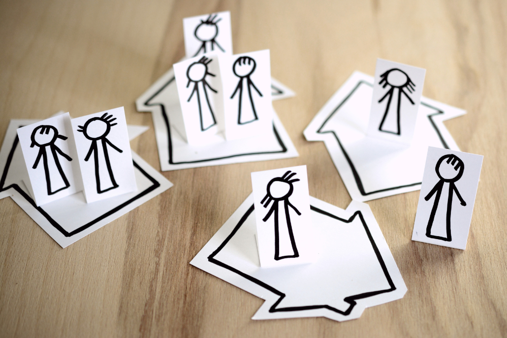 A photo of stick drawing of people standing on cut-out drawings of houses. 