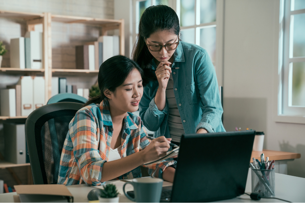 Two women looking at a computer. One woman is sitting down at and is pointing to a notebook with a pen. The other woman is standing up.