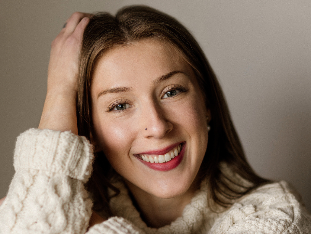 A headshot of Katie with shoulder length brown hair smiling, wearing a white sweater.