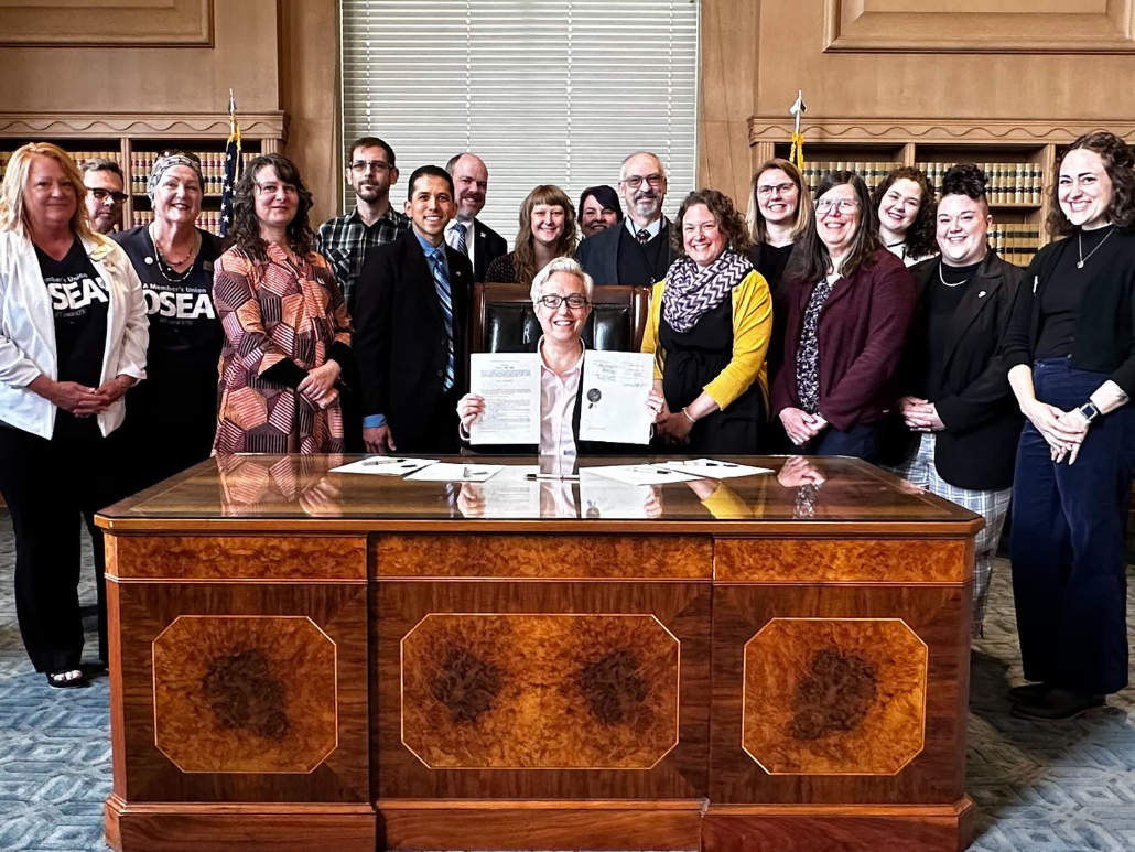 A photo of the Oregon Governor at a desk surrounded by people after signing legislation into law.