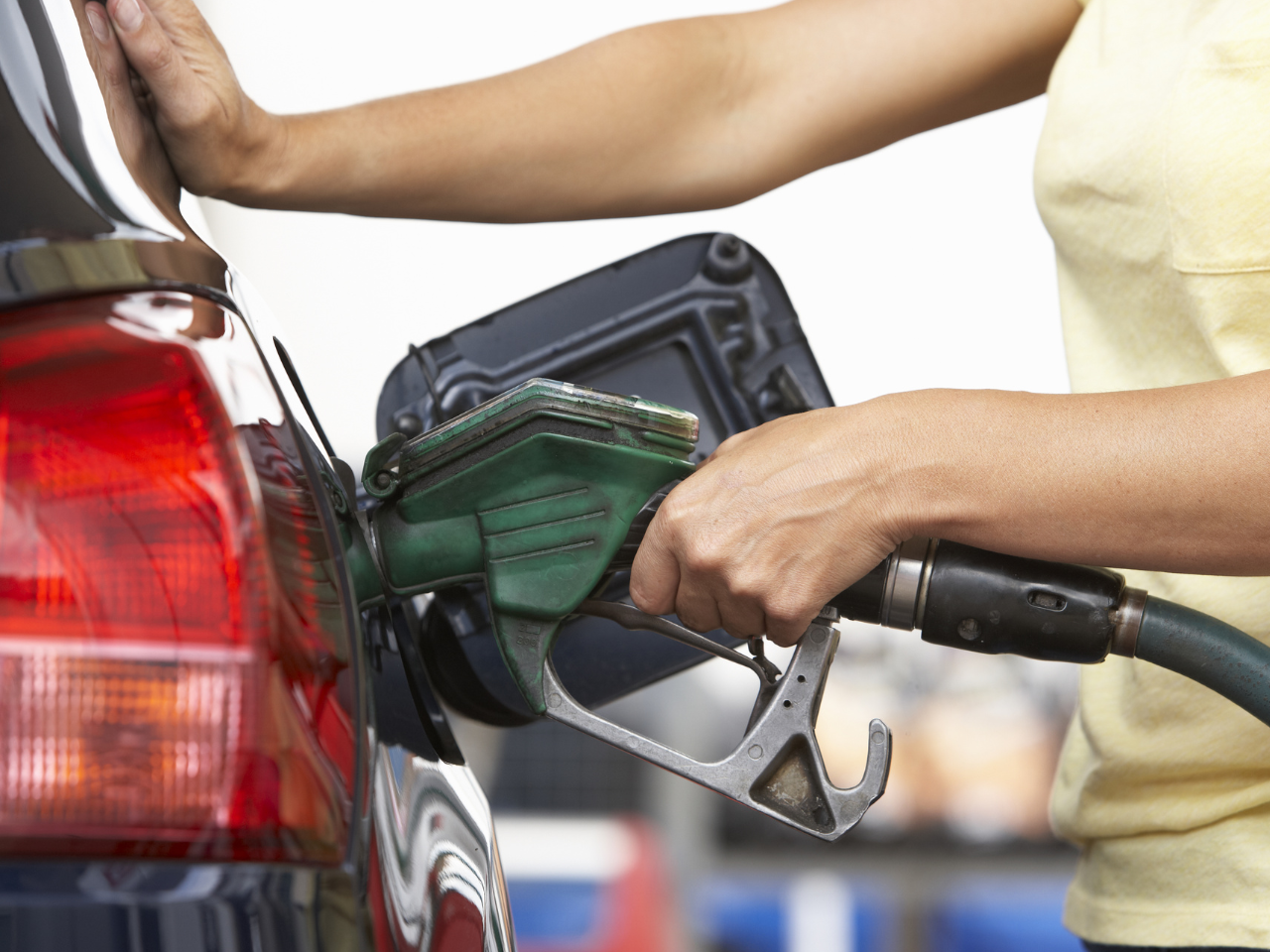 Close-up of a person refueling a car, holding a green fuel nozzle and pressing the car with the other hand.