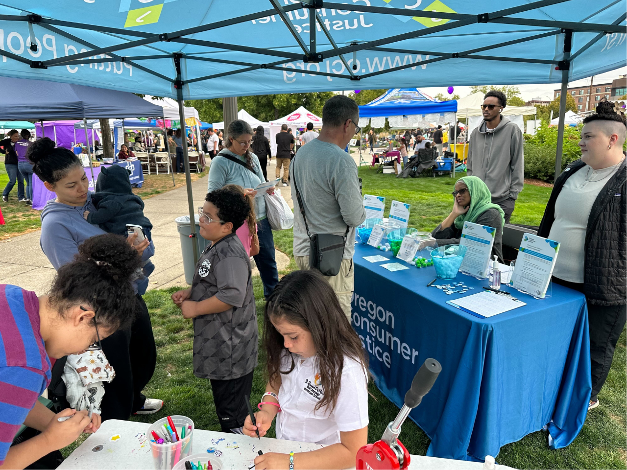 Evento Viva Salem 2023 con personas reunidas alrededor de un stand debajo de una carpa con la etiqueta "Oregon Consumer Justice", niños y adultos interactuando, y materiales informativos sobre la mesa.