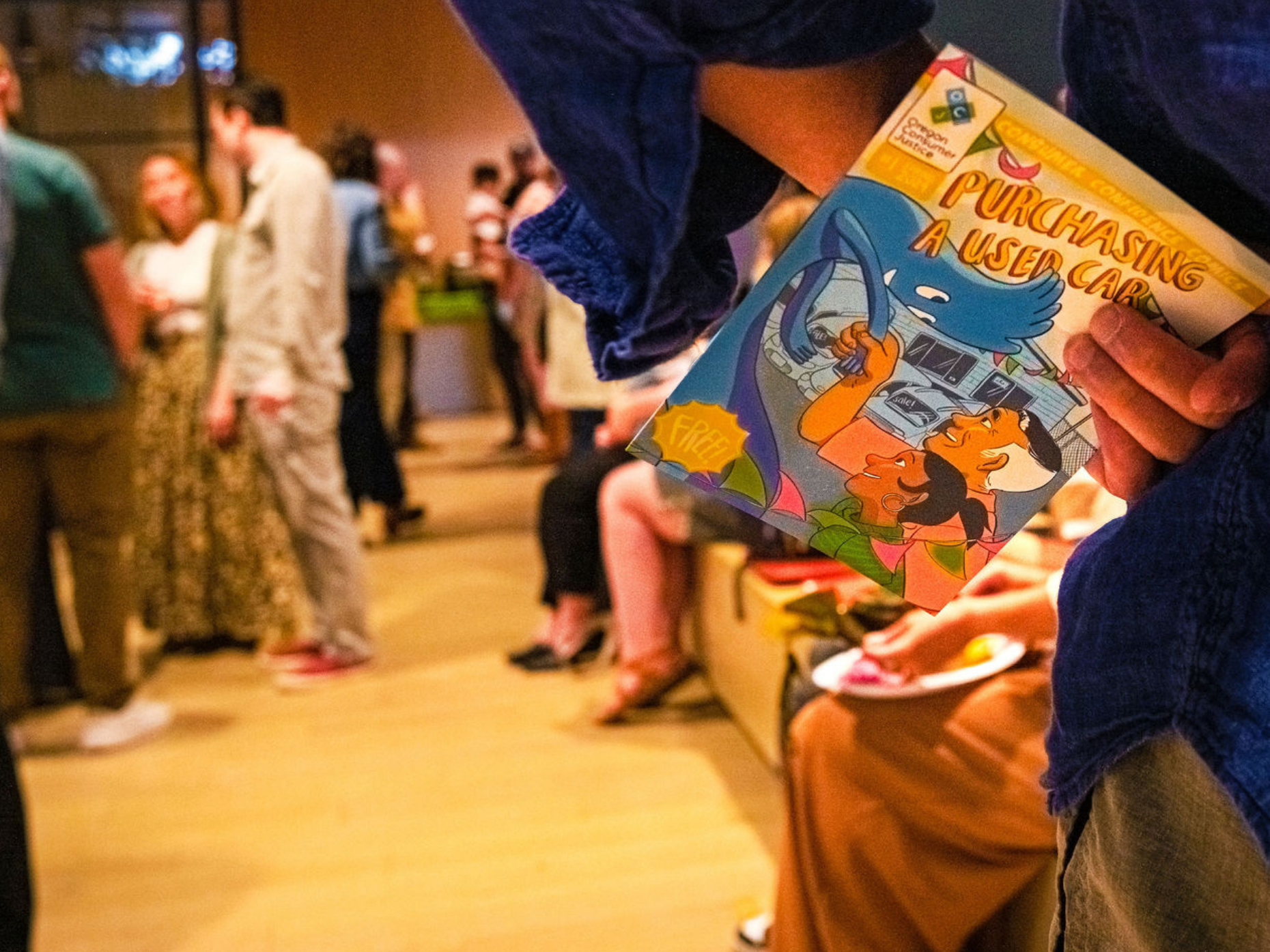 A person holding a comic book titled 'Purchasing a Used Car' at a social event with people in the background.