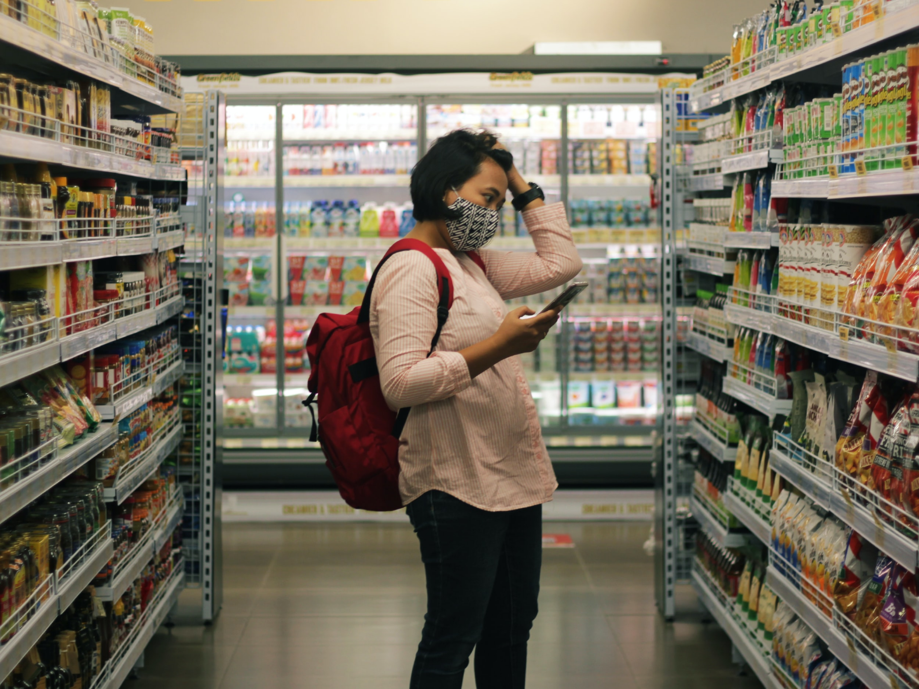 Woman in a grocery store aisle wearing a mask and using her phone.