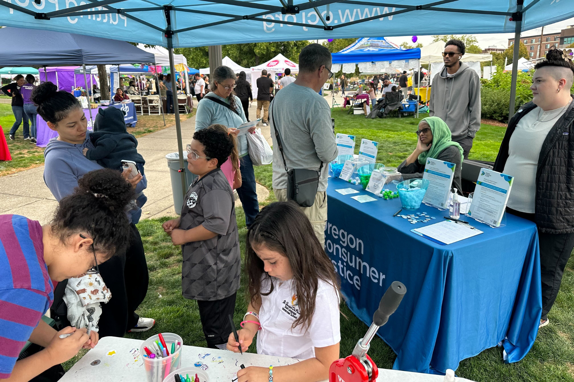 2023 Viva Salem event with people gathered around a booth under a tent labeled "Oregon Consumer Justice," children and adults interacting, and informational materials on the table.