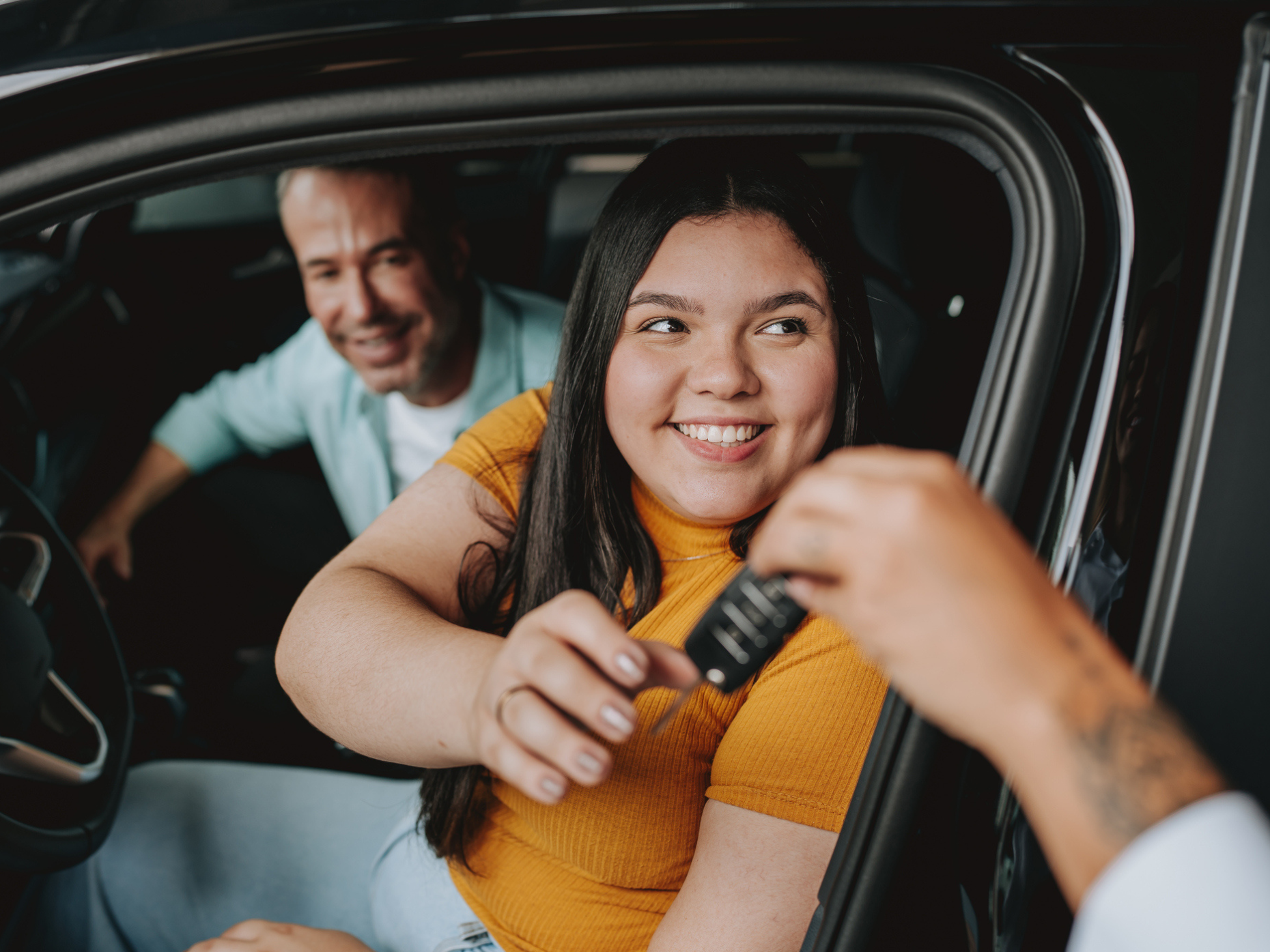 Woman smiling seated in the driver’s seat of a car as she takes the keys from a person out of frame. There is a man in the passenger seat, also smiling. 