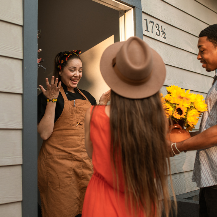 A couple standing at the doorstep of a house, presenting a bouquet of sunflowers to a smiling woman in an apron.