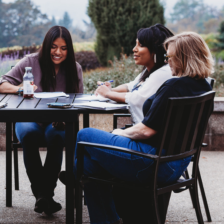 Three women sitting at a table outdoors, smiling and working on documents together.