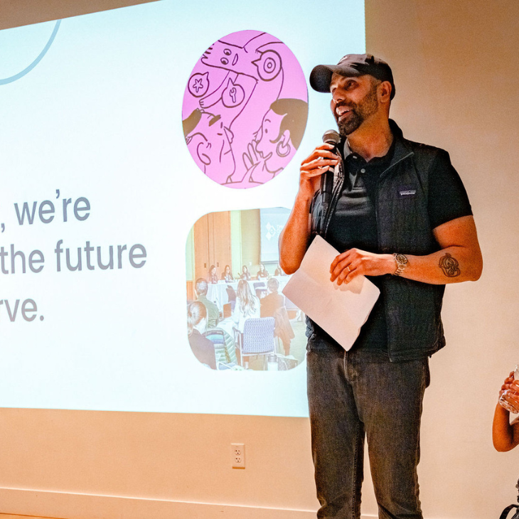 OCJ Executive Director, Jagjit Nagra holds a mic as he addresses a group of people at an event with the OCJ logo and writing on a screen that reads “Building the future we deserve.”