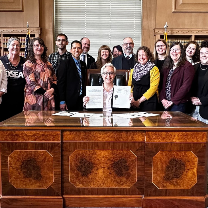 A group of advocates standing behind a large wooden desk, with Governor Tina Kotek, seated in the center holding up the ceremonially signed documents for SB 1595. They are in a formal room with flags and bookshelves in the background.