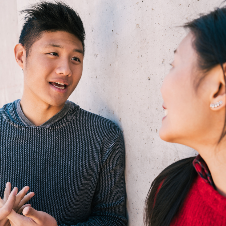 A young man is leaning against a light-colored outdoor wall speaking to a person who is just out of frame. 