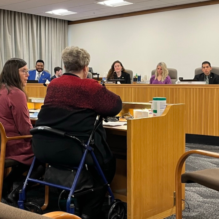 A legislative meeting room with a panel of Oregon House Representatives sitting at a long desk, facing a woman using a walker who is testifying on behalf of a bill to change debt collection practices in Oregon with OCJ’s policy director seated next to her. 