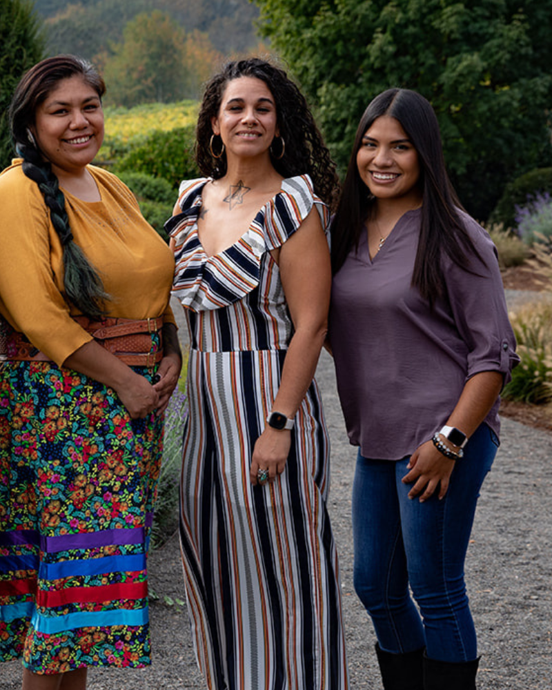 Three members of OCJ’s inaugural Community Cohort standing outdoors, smiling at the camera. One wears a yellow top, one a striped dress, and one a purple blouse.