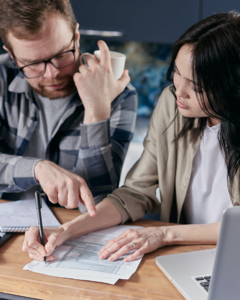 A man and woman sitting at a table with a laptop, calculator, and documents, discussing financial matters.