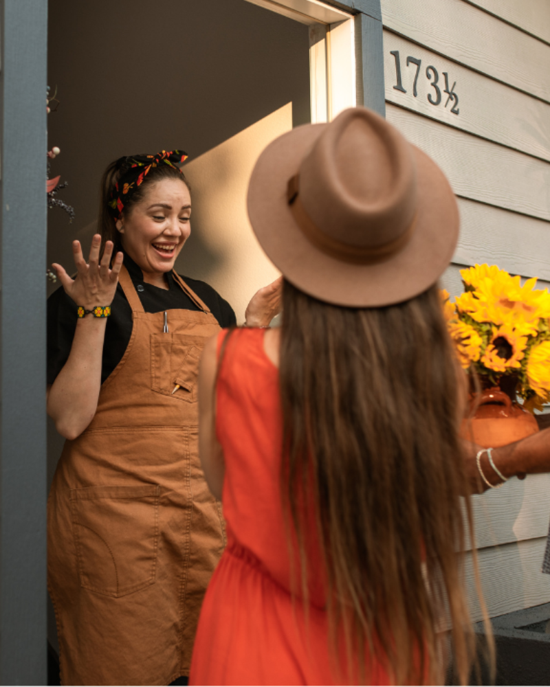 A couple standing at the doorstep of a house, presenting a bouquet of sunflowers to a smiling woman in an apron.