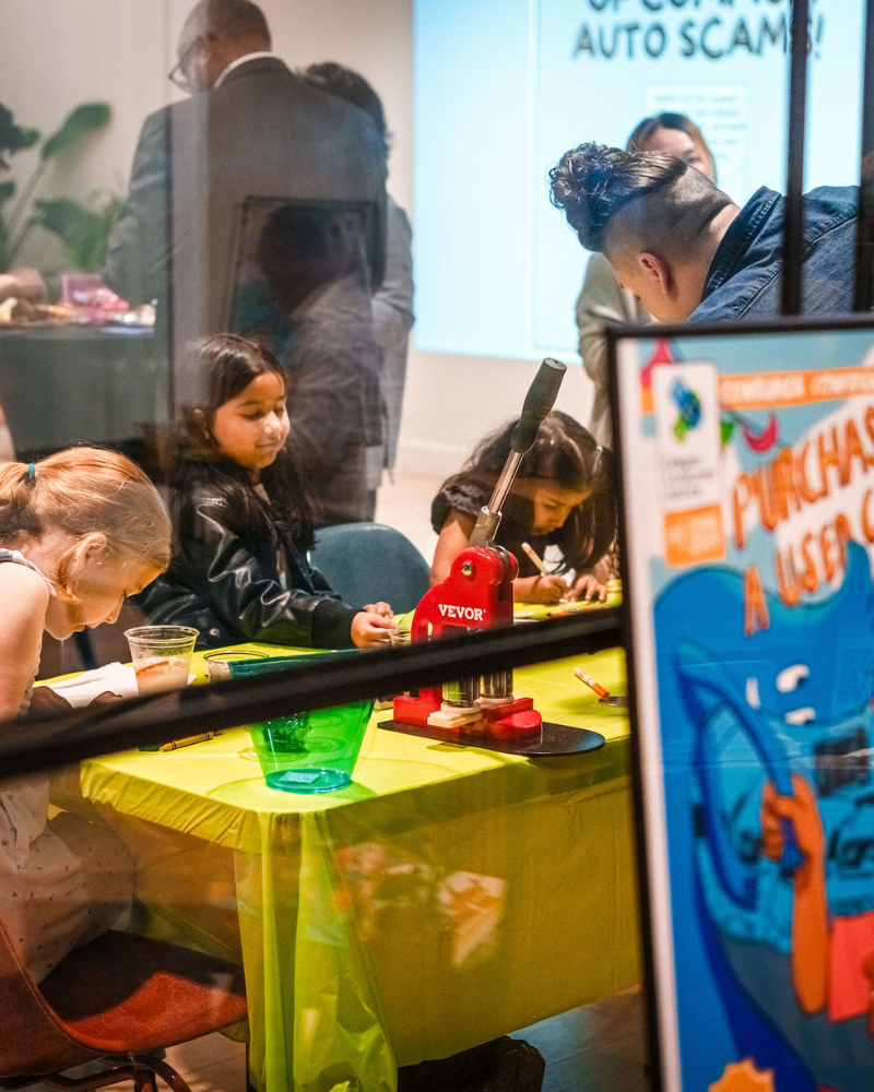 A group of children and adults are engaged in an activity at a community event, with a colorful poster titled "Purchasing a Used Car" visible in the foreground.