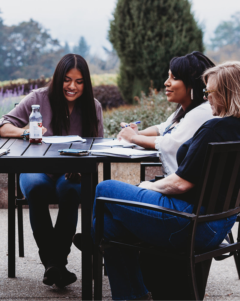 Three women sitting at a table outdoors, smiling and working on documents together.