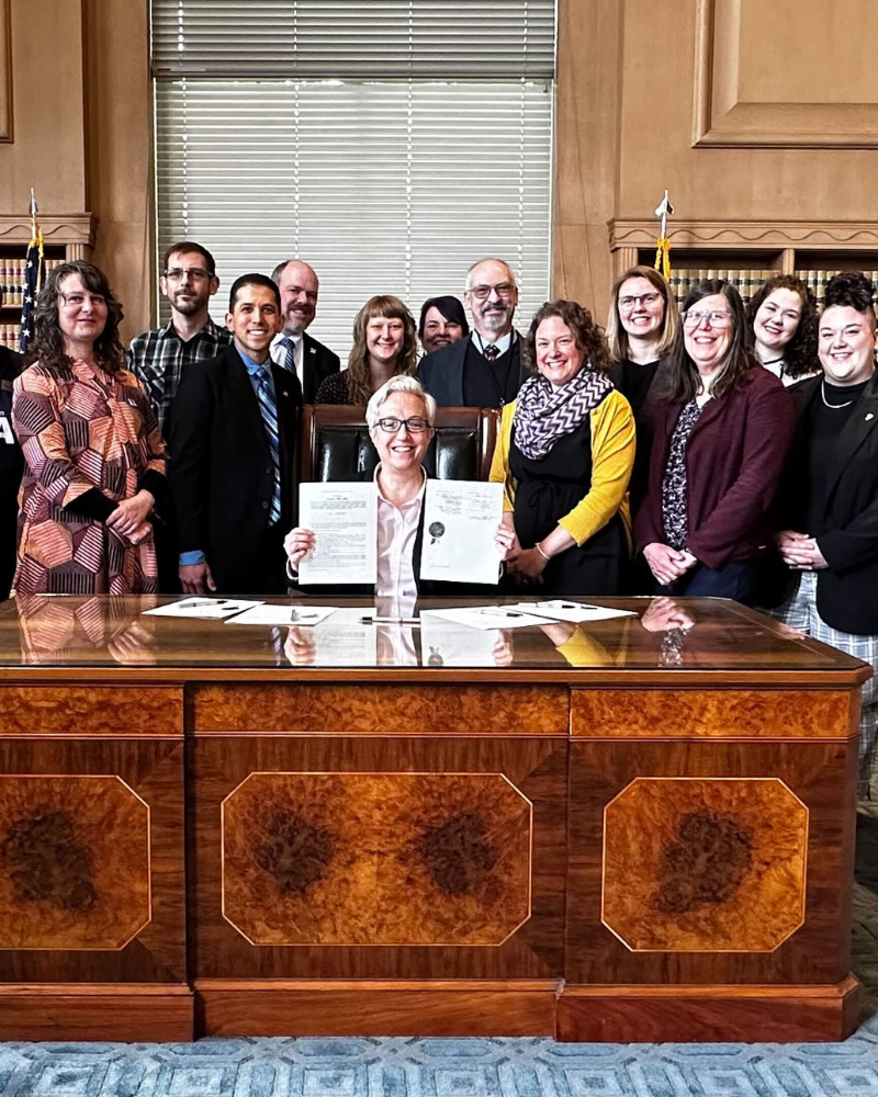  A group of advocates standing behind a large wooden desk, with Governor Tina Kotek, seated in the center holding up the ceremonially signed documents for SB 1595. They are in a formal room with flags and bookshelves in the background.