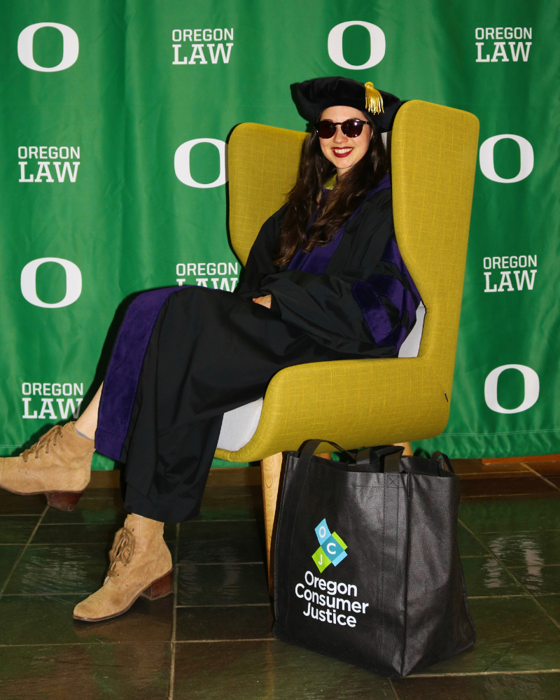 Former intern, Taylor Westlund, in her graduation robes and a cap, wearing sunglasses, sitting in a yellow chair with an UO Law backdrop. There is a black Oregon Consumer Justice tote bag on the floor next to her.