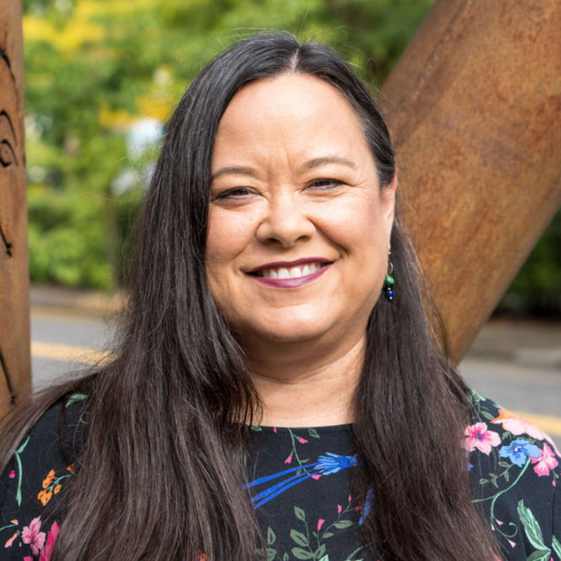 A woman with long dark hair, smiling, wearing a black top with floral patterns, standing outdoors with a wooden structure in the background.