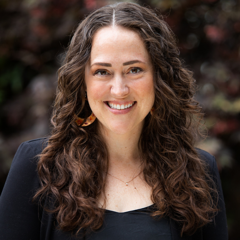 A woman with long curly brown hair, smiling, wearing a black top, standing in front of a background with colorful foliage.