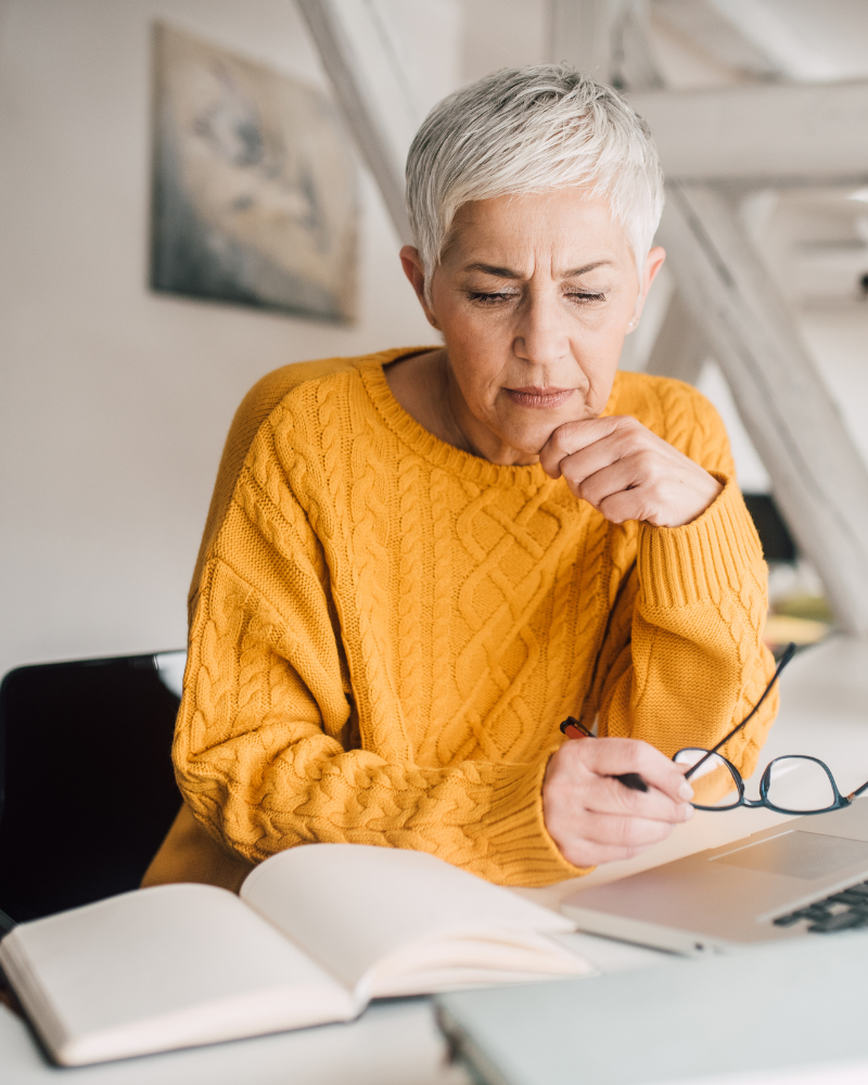 Woman with short white hair wearing a yellow sweater, sitting and looking at papers on a desk.