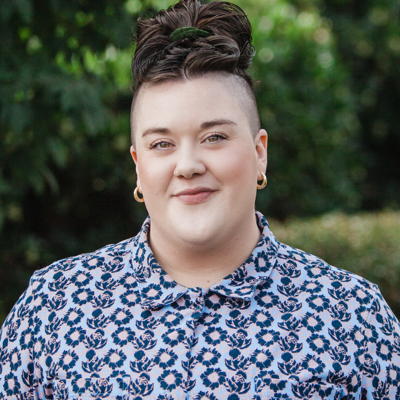 Person in a patterned blue collared shirt with brown hair in a high bun smiling