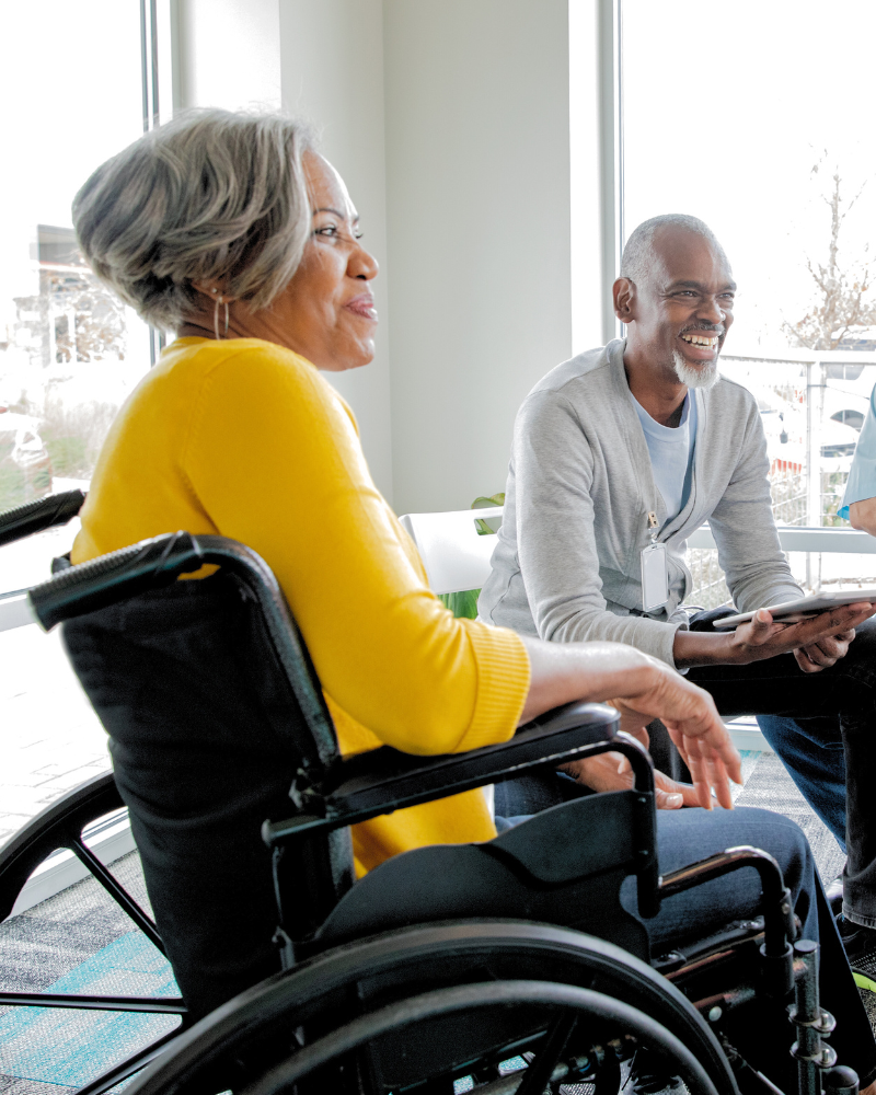 An older woman in a wheelchair next to a man sitting on a couch. Both are smiling.