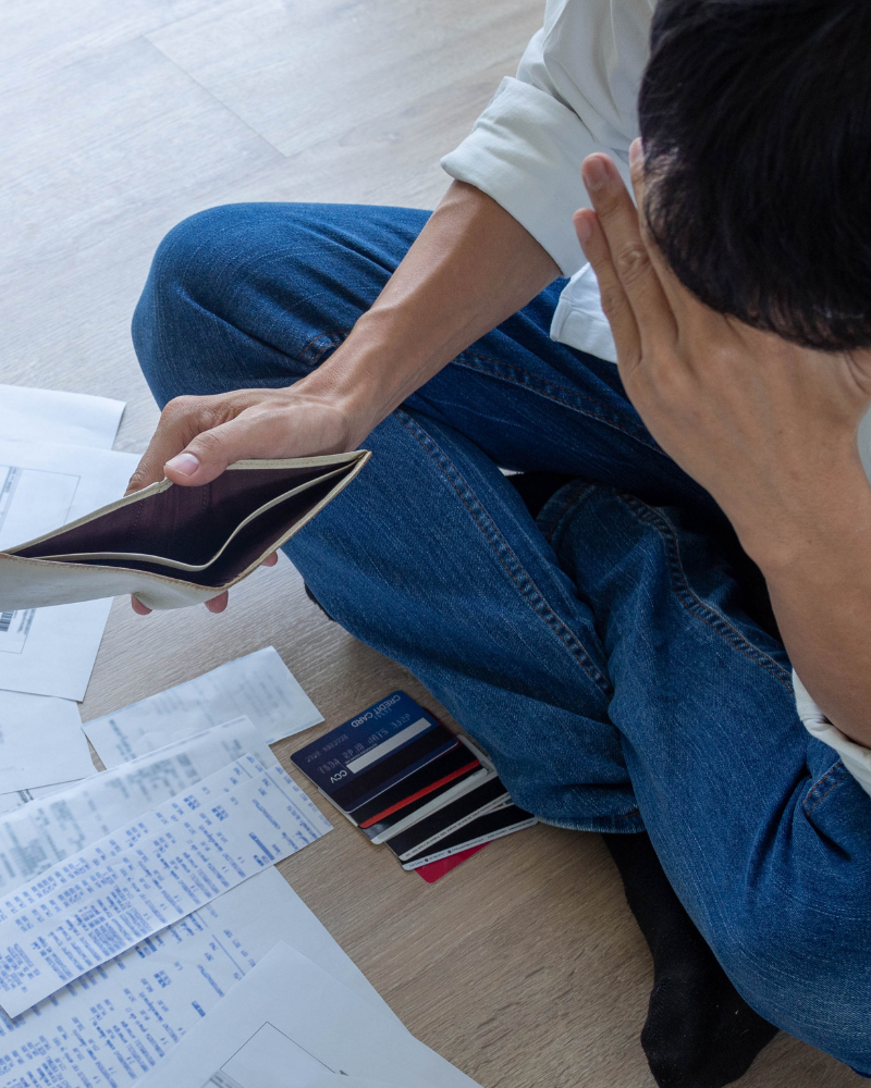 A person sitting on the floor in front of papers and credit cards. They are holding their head in one hand and an empty wallet in the other.