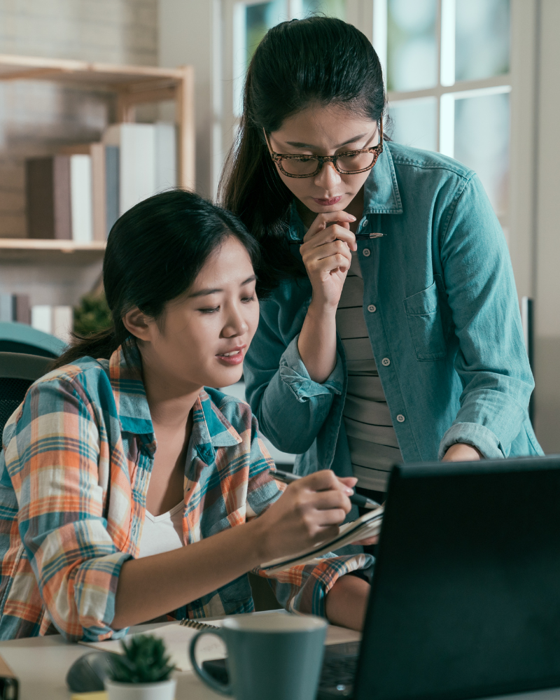 Two women looking at a computer. One woman is sitting down at and is pointing to a notebook with a pen. The other woman is standing up.